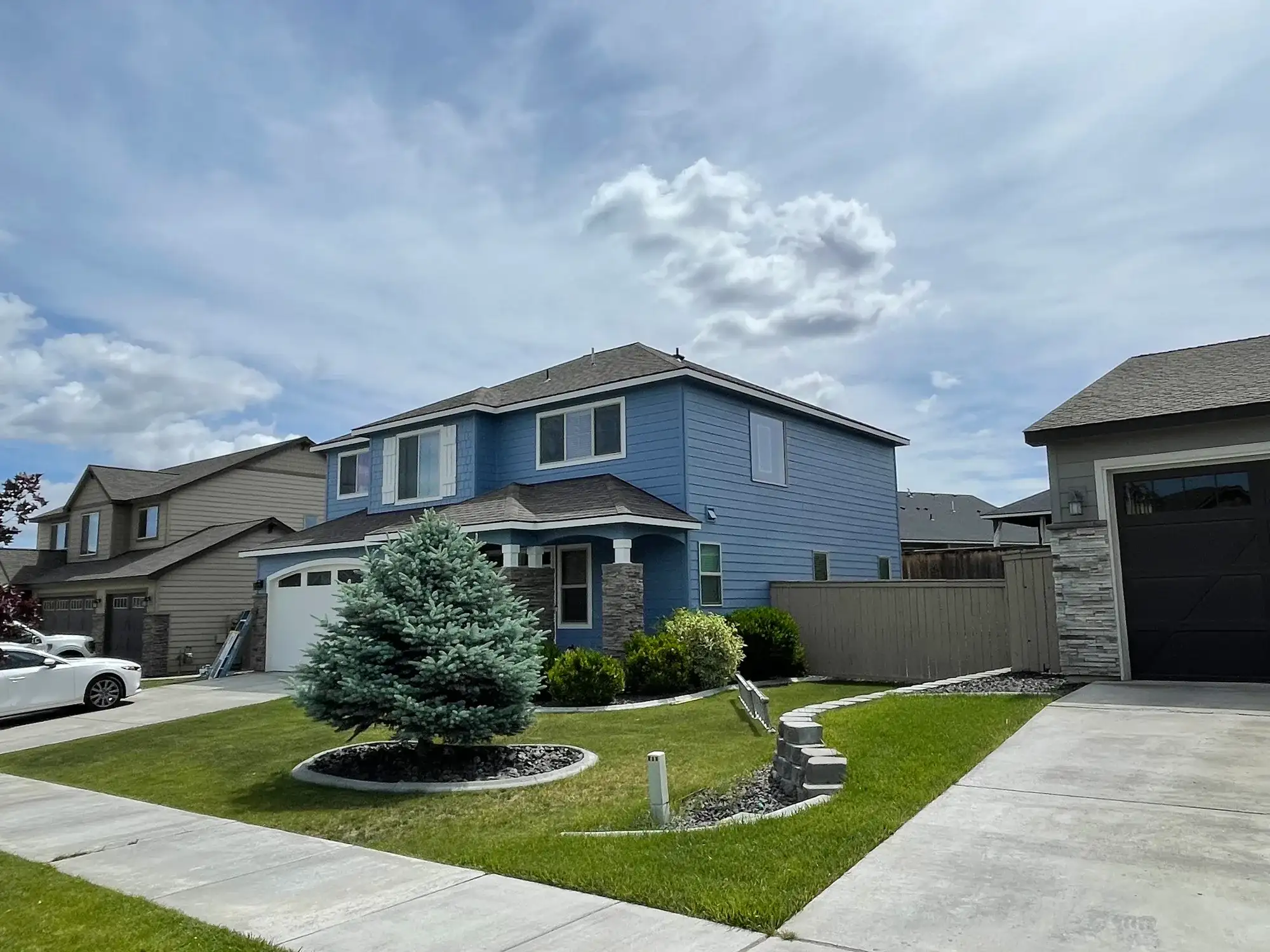 Front of a two-story home painted blue with a white garage door, stone columns and a blue spruce in the yard