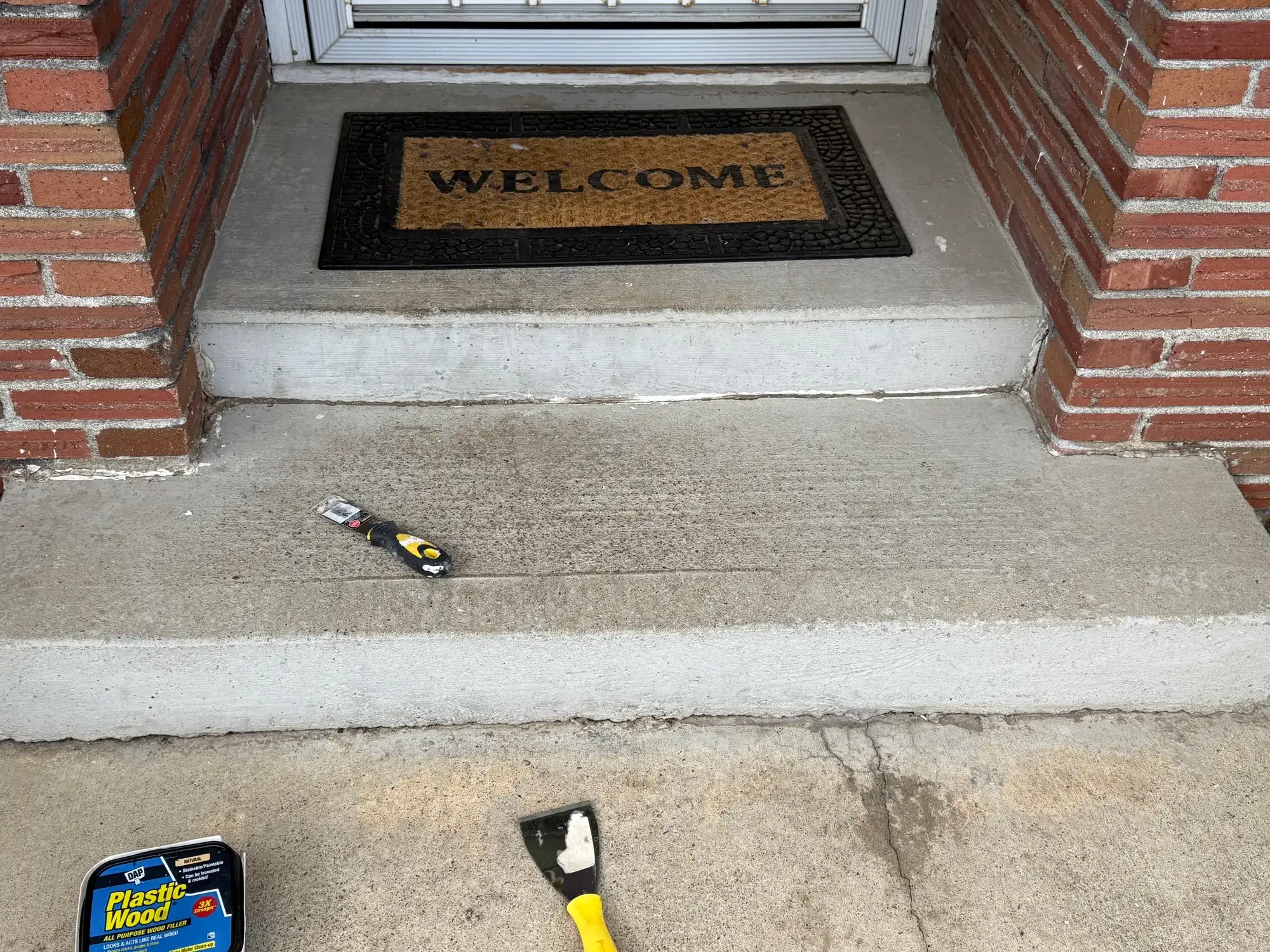 Concrete front step being patched, with a scraper and wood filler on the step